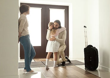 A girl hugging her dad, affected by interstate parenting plans, while her mother stands by a nearby wall.